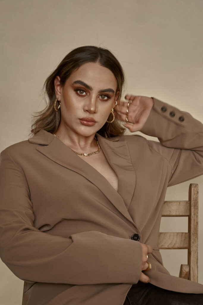Valeria Cisneros a model with striking features poses elegantly in a beige blazer gold jewelry and dark pants against a muted backdrop.